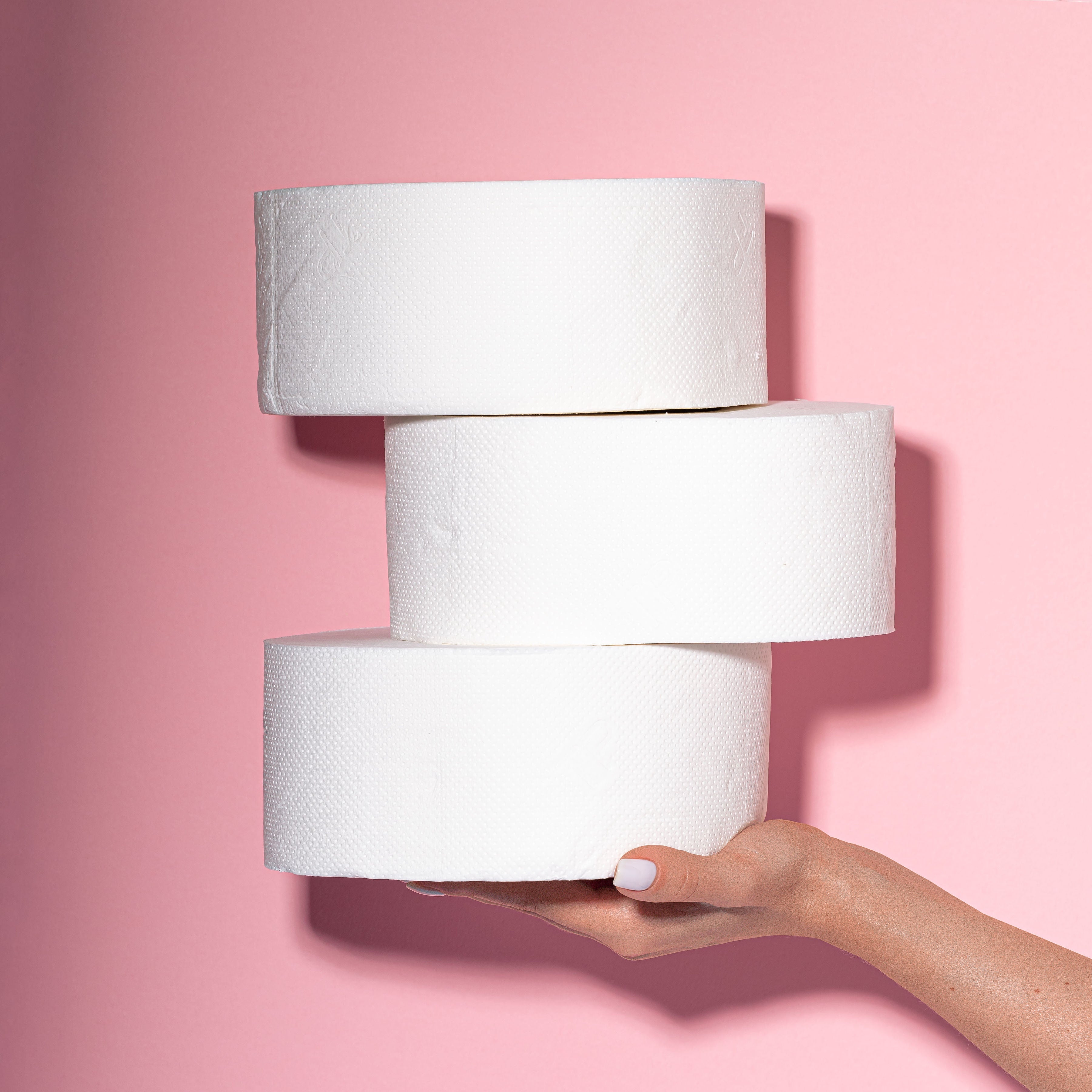 Hand holding three rolls of jumbo toilet paper against a pink background